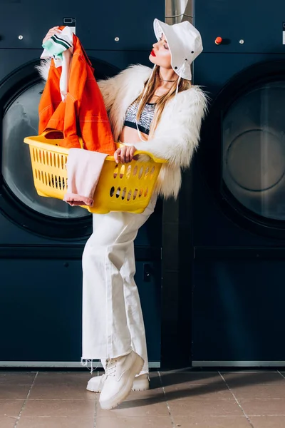 Élégante jeune femme en fausse veste de fourrure et chapeau tenant panier et regardant la blanchisserie près des machines à laver dans la laverie automatique — Photo de stock