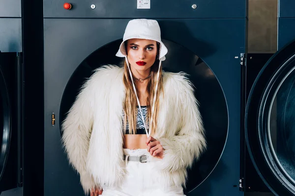 Stylish young woman in faux fur jacket and hat near washing machines in laundromat — Stock Photo