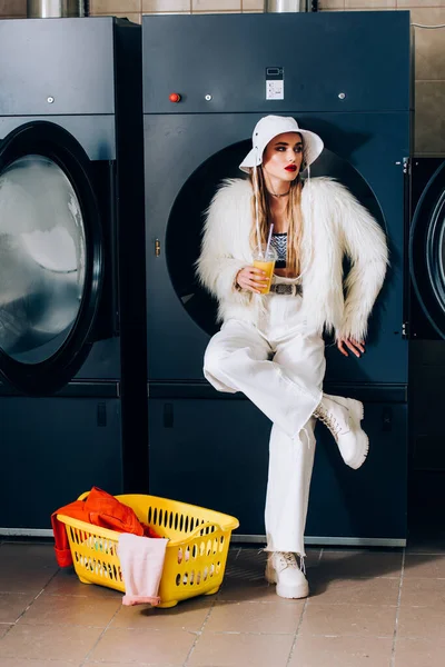 Femme élégante en fausse veste de fourrure et chapeau tenant tasse en plastique avec jus d'orange près du panier avec blanchisserie et lave-linge dans la laverie automatique — Photo de stock