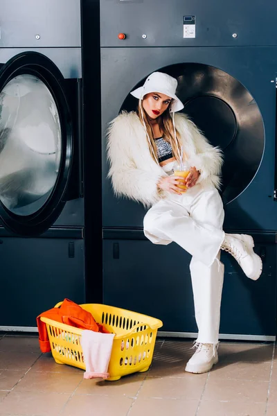 Mujer joven en chaqueta de piel sintética y sombrero sosteniendo taza de plástico con zumo de naranja cerca de la cesta con lavanderia y lavadoras en lavandería - foto de stock