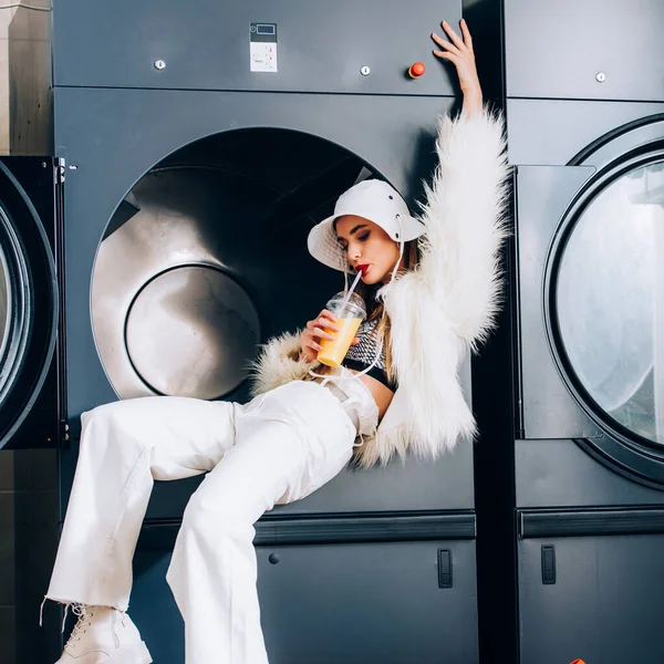 Модна молода жінка в штучній шубці і капелюсі тримає пластикову чашку і п'є апельсиновий сік біля пральних машин в пральному апараті — стокове фото