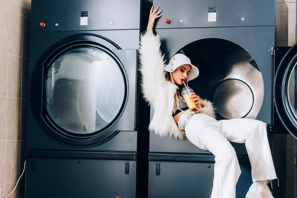 Trendfrau in Kunstpelzjacke und Hut hält Plastikbecher in der Hand und trinkt Orangensaft neben Waschmaschinen im öffentlichen Waschsalon — Stockfoto