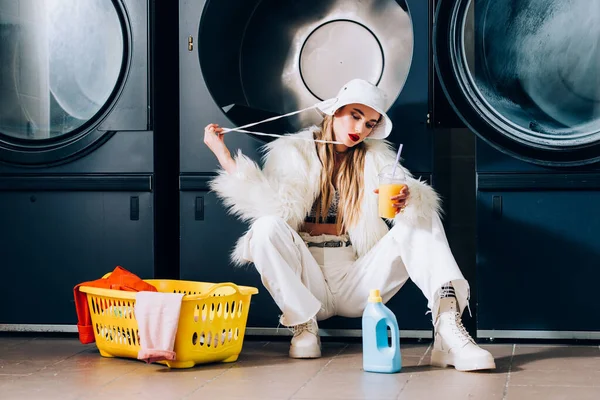 Stylish woman in faux fur jacket and hat holding plastic cup with orange juice near basket with laundry and washing machines in laundromat — Stock Photo