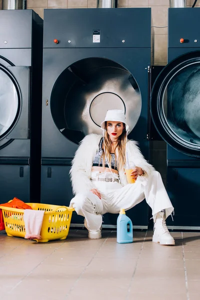 Stylish woman in faux fur jacket and hat holding plastic cup with orange juice near basket with laundry and washing machines in laundromat — Stock Photo