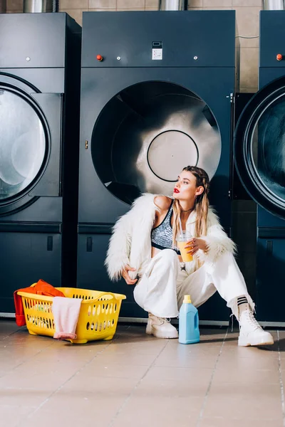 Stylish woman in faux fur jacket holding plastic cup with orange juice near basket with laundry and detergent bottle in laundromat — Stock Photo