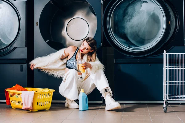 Young woman in faux fur jacket holding plastic cup with orange juice near basket with laundry, detergent bottle and washing machines in laundromat — Stock Photo