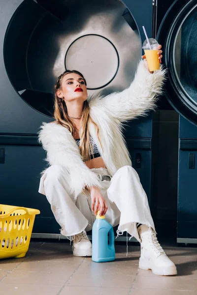 Jovem elegante em casaco de pele falso segurando copo de plástico com suco de laranja perto cesta com lavanderia, garrafa de detergente e máquinas de lavar roupa — Fotografia de Stock