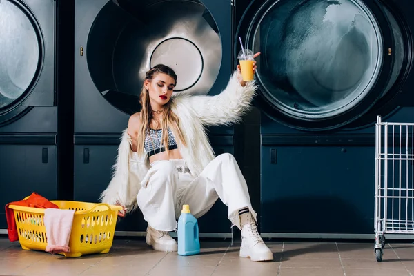 Donna in giacca di pelliccia sintetica contenente bicchiere di plastica con succo d'arancia vicino al cestino con lavanderia, bottiglia detergente e lavatrici in lavanderia — Foto stock