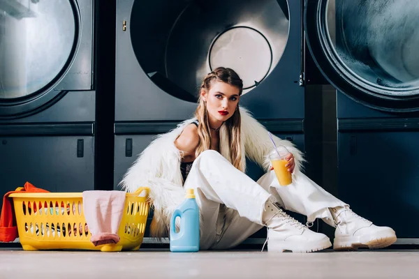 Стильная женщина в искусственной меховой куртке с пластиковой чашкой с апельсиновым соком возле корзины со стиркой, бутылкой моющего средства и стиральными машинами в прачечной — стоковое фото