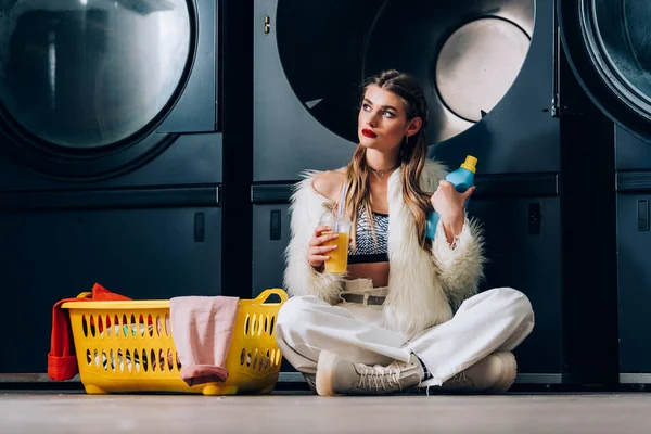 Mulher elegante em casaco de pele falso segurando copo de plástico com suco de laranja perto da cesta com lavanderia, garrafa de detergente e máquinas de lavar roupa na lavanderia — Fotografia de Stock