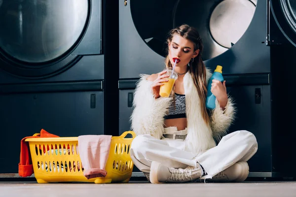 Mulher elegante em casaco de pele falso segurando copo de plástico com suco de laranja perto da cesta com lavanderia, garrafa de detergente e máquinas de lavar roupa na lavanderia — Fotografia de Stock