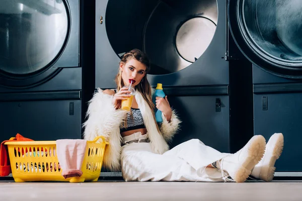 Stylish woman in faux fur jacket drinking orange juice and sitting near basket with laundry, detergent bottle and washing machines in laundromat — Stock Photo