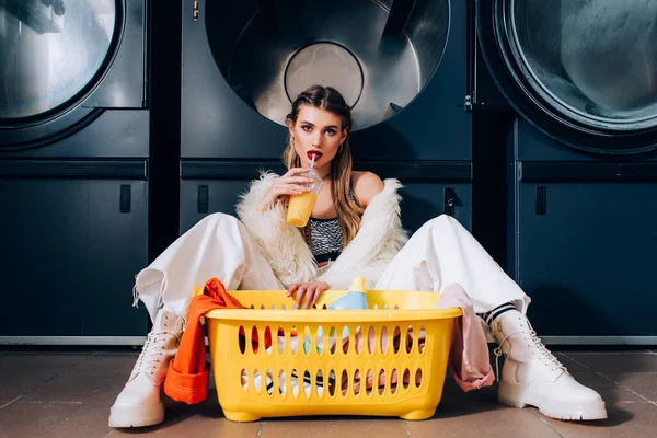 Woman in faux fur jacket drinking orange juice and sitting near basket with laundry, detergent bottle and washing machines in laundromat — Stock Photo