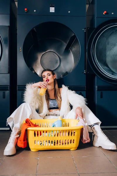 Junge Frau in Kunstpelzjacke trinkt Orangensaft und sitzt neben Korb mit Wäsche, Waschmittelflasche und Waschmaschinen im Waschsalon — Stockfoto