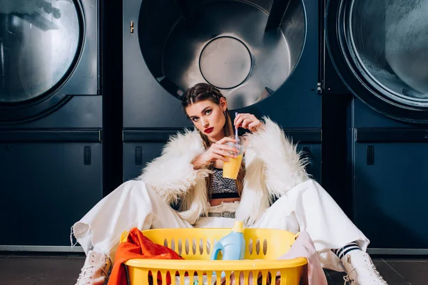 Young woman holding plastic cup with orange juice and sitting near basket with laundry, detergent bottle and washing machines in laundromat — Stock Photo
