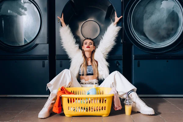 Young woman in faux fur jacket sitting with hands above head and looking at camera in laundromat — Stock Photo