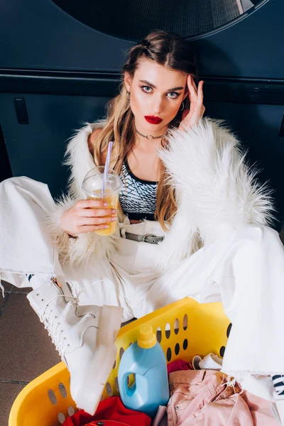 Young woman in faux fur jacket sitting with orange juice near basket with dirty clothing — Stock Photo