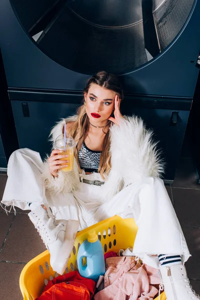 Jovem mulher em casaco de pele falso sentado com suco de laranja perto cesta com roupas sujas na lavanderia — Fotografia de Stock