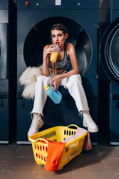 Stylish woman sitting in washing machine near faux fur jacket and drinking orange juice near basket with dirty clothing in laundromat — Stock Photo