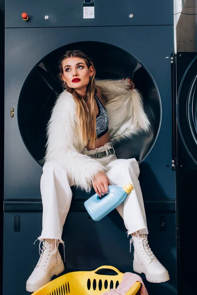 Jeune femme en fausse fourrure blanche assise dans la machine à laver et tenant bouteille avec détergent dans la laverie automatique — Photo de stock