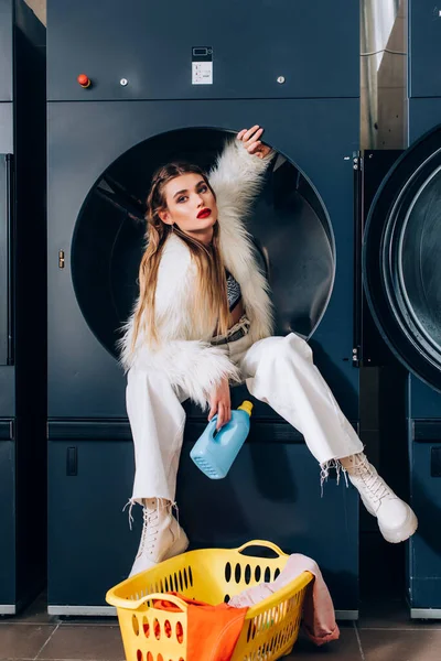 Trendy woman in white faux fur jacket sitting in washing machine and holding bottle with detergent in laundromat — Stock Photo