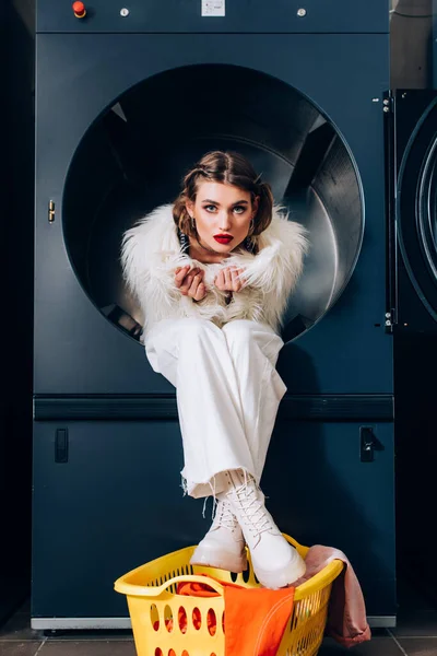 Модна жінка в білій хутряній куртці, що сидить у пральній машині біля одягу в кошику — стокове фото