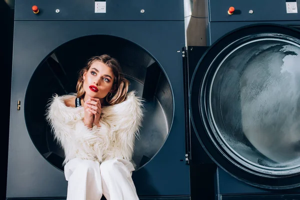 Мрійлива жінка в білій хутряній куртці, що сидить у громадській пральній машині — стокове фото