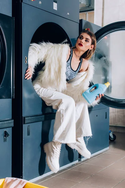 Молода стильна жінка тримає пляшку з миючим засобом і сидить у громадській пральній машині — стокове фото