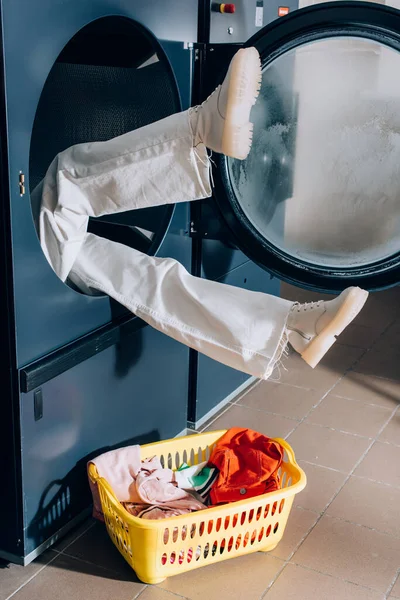Ноги женщины торчат из стиральной машины возле корзины с грязным бельем — стоковое фото
