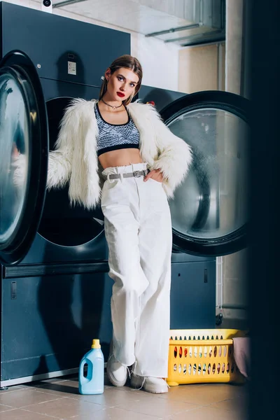 Junge stylische Frau in Kunstpelzjacke, die mit der Hand in der Tasche im öffentlichen Waschsalon steht — Stockfoto