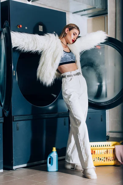 Giovane donna elegante in pelliccia sintetica giacca in piedi vicino cesto e bottiglia con detergente in lavanderia pubblica — Foto stock