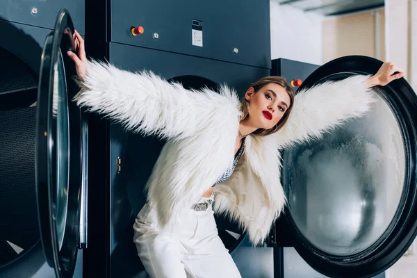 Jovem mulher na moda em casaco de pele falso em pé perto de máquinas de lavar roupa na lavanderia pública — Fotografia de Stock