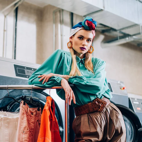 Mujer joven y elegante en turbante de pie cerca de perchero de ropa en lavandería - foto de stock