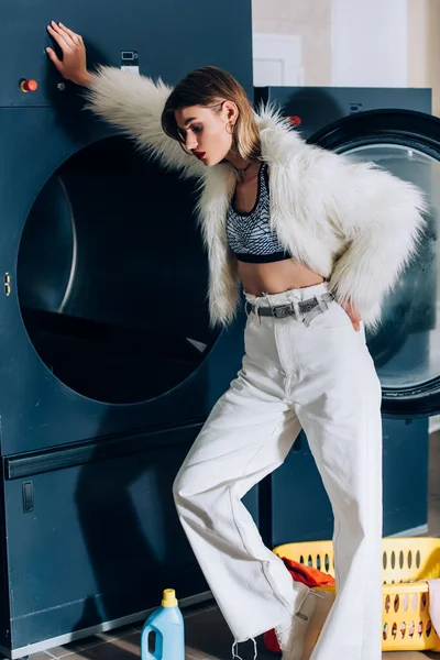 Young stylish woman in faux fur jacket leaning on washing machine in public laundromat — Stock Photo