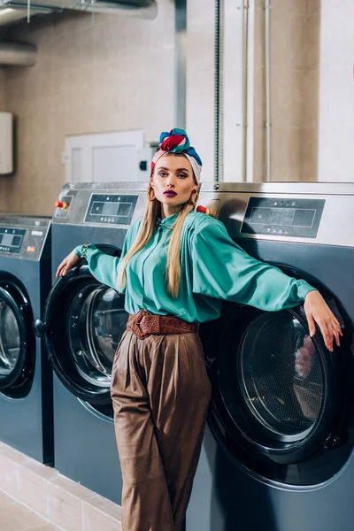 Stylish woman in turban standing near washing machines in laundromat — Stock Photo