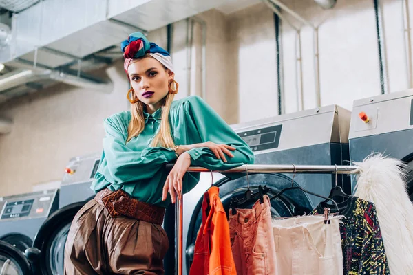 Jeune femme élégante en turban debout près des machines à laver dans la laverie automatique — Photo de stock