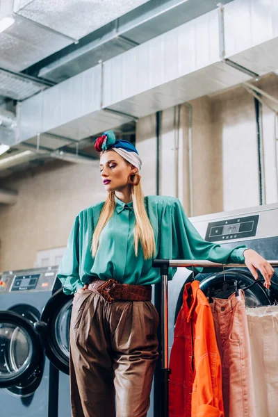 Stylish woman in turban standing near washing machines and clothing in laundromat — Stock Photo