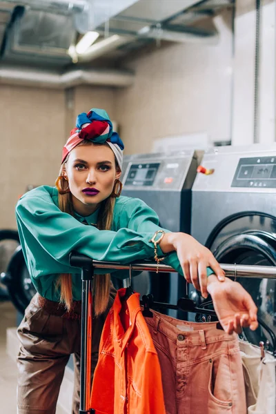 Stylische Frau im Turban steht neben Kleidung auf Kleiderbügeln und Waschmaschinen im Waschsalon — Stockfoto