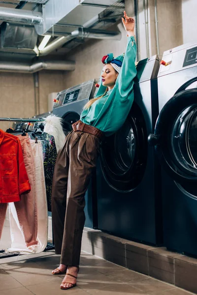 Femme à la mode en turban debout près du porte-vêtements et des machines à laver dans la laverie automatique — Photo de stock