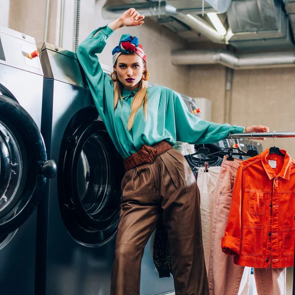 Молодая модная женщина в тюрбане стоит рядом с вешалкой для одежды и стиральными машинами в современной прачечной — стоковое фото