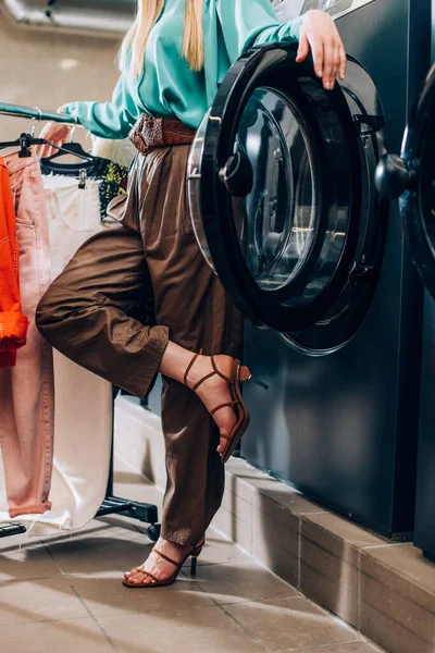 Vista recortada de la mujer con estilo de pie cerca de perchero de ropa y lavadora en la lavandería moderna - foto de stock