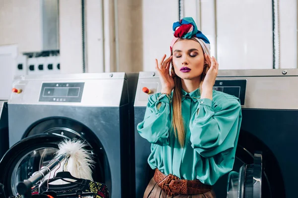 Stylish woman touching turban and standing near washing machines in modern laundromat — Stock Photo