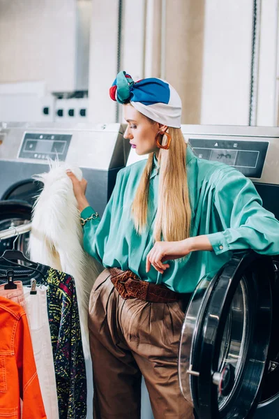 Mulher elegante em turbante de pé perto de rack de roupas e máquinas de lavar roupa na lavanderia moderna — Fotografia de Stock