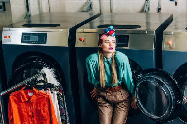 Young woman in turban standing near washing machines and clothing rack in laundromat — Stock Photo