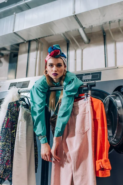 Молодая женщина в тюрбане опирается на вешалку для одежды и стиральные машины в прачечной — стоковое фото