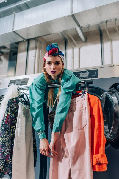 Jeune femme en turban appuyé sur le porte-vêtements et les machines à laver dans la laverie automatique — Photo de stock