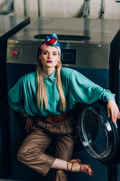 Stylish woman in turban standing near washing machine in laundromat — Stock Photo