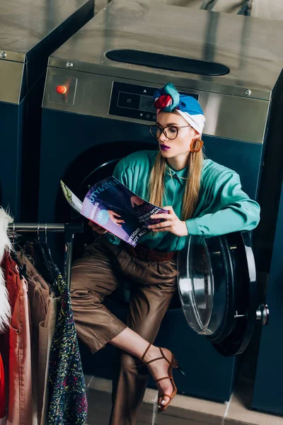 Jeune et élégante femme en turban et lunettes de lecture magazine en laverie automatique — Photo de stock
