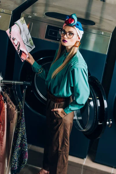 Young and stylish woman in turban and glasses holding magazine and standing with hand in pocket in laundromat — Stock Photo