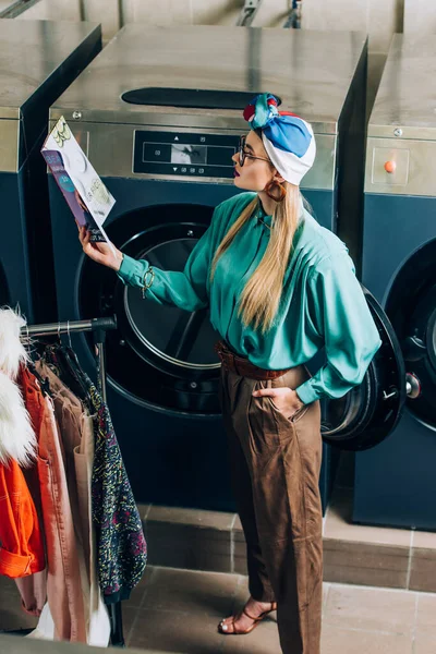 Mujer con estilo en turbante y gafas de lectura revista y de pie con la mano en el bolsillo en la lavandería - foto de stock